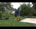 Jill wearing a shiny yellow rainpants and rain jacket pulls over a raincoat and playing with water our of the water hose (Video)