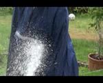 Jill wearing a shiny yellow rainpants and rain jacket pulls over a raincoat and playing with water our of the water hose (Video)