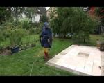 Jill wearing a shiny yellow rainpants and rain jacket pulls over a raincoat and playing with water our of the water hose (Video)