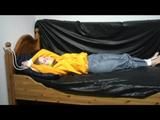 Samantha in a shiny yellow rain jacket and a shiny grey rain trowsers tied and gagged on bed (Video)
