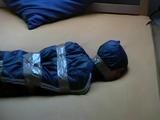 One of our archive girl tied, hooded and gagged by tape on a bed wearing shiny nylon rainwear (Video)