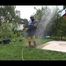 Jill wearing a shiny yellow rainpants and rain jacket pulls over a raincoat and playing with water our of the water hose (Video)