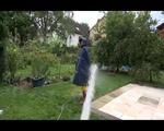 Jill wearing a shiny yellow rainpants and rain jacket pulls over a raincoat and playing with water our of the water hose (Video)