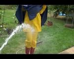 Jill wearing a shiny yellow rainpants and rain jacket pulls over a raincoat and playing with water our of the water hose (Video)