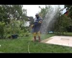 Jill wearing a shiny yellow rainpants and rain jacket pulls over a raincoat and playing with water our of the water hose (Video)