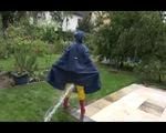 Jill wearing a shiny yellow rainpants and rain jacket pulls over a raincoat and playing with water our of the water hose (Video)